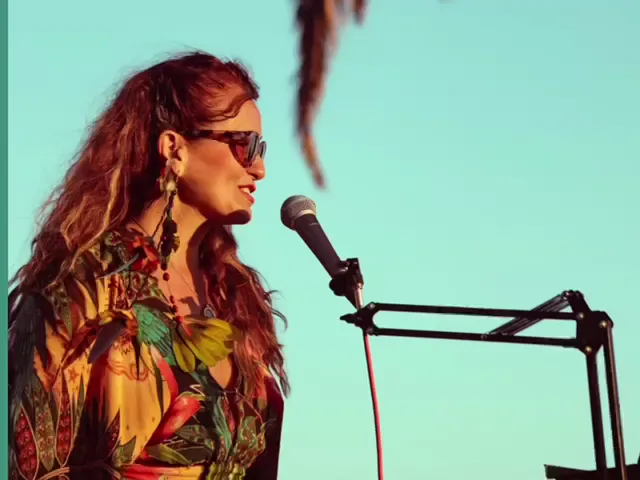 Vanessa Falabella singing into a microphone in profile, against a clear sky